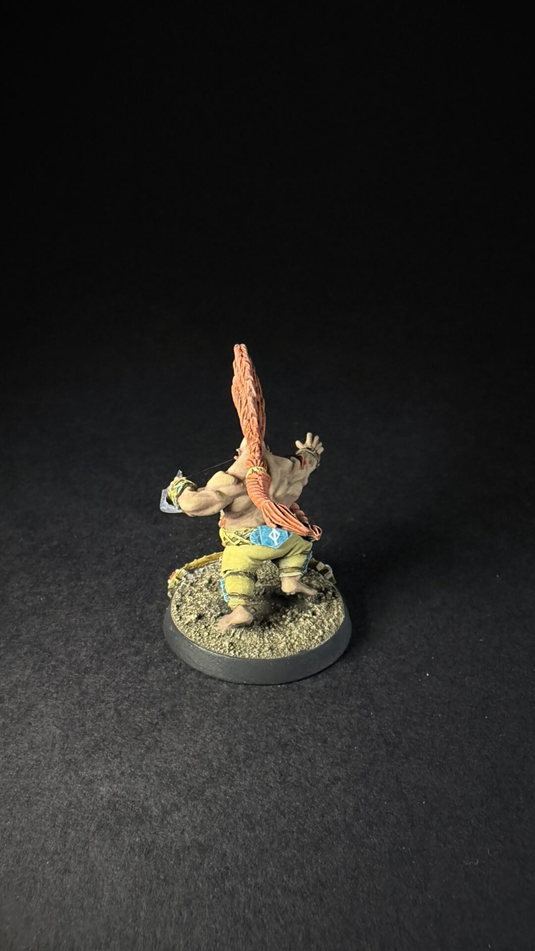 Back view of a painted tabletop miniature: muscular humanoid with long orange hair, wearing green pants, standing on a rocky base.