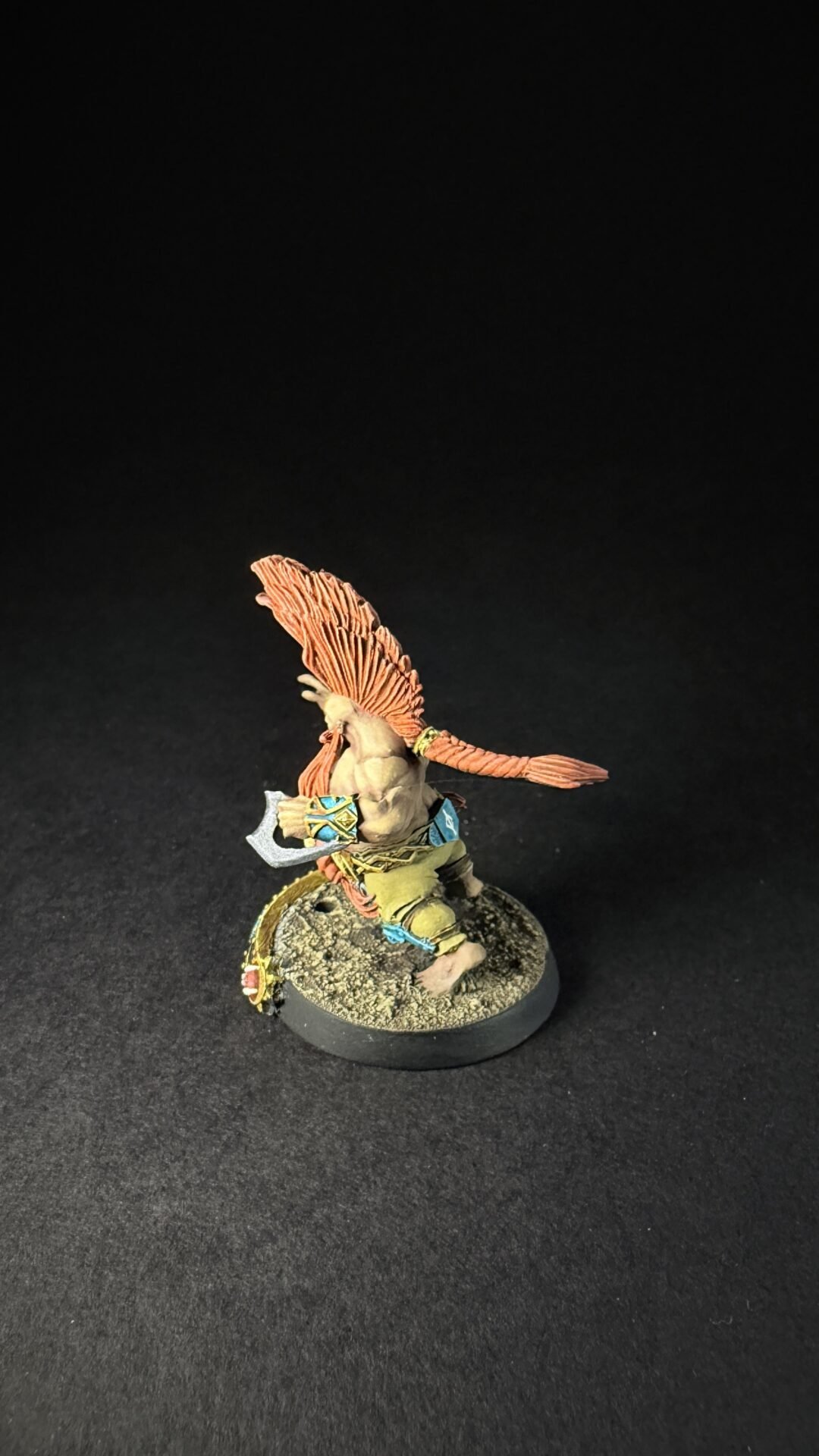 Small fantasy warrior miniature with an orange feathered headdress wielding a small axe, standing on a rocky base.