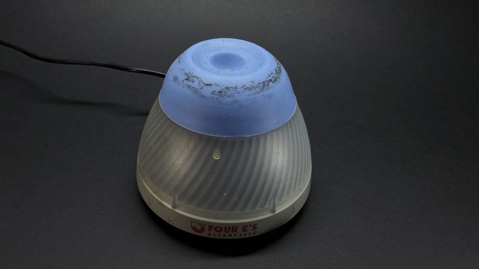 Blue, dome-shaped lab light with a ribbed white base sits on a dark surface; power cord extends to the left and the Four E's Scientific logo is visible on the base.