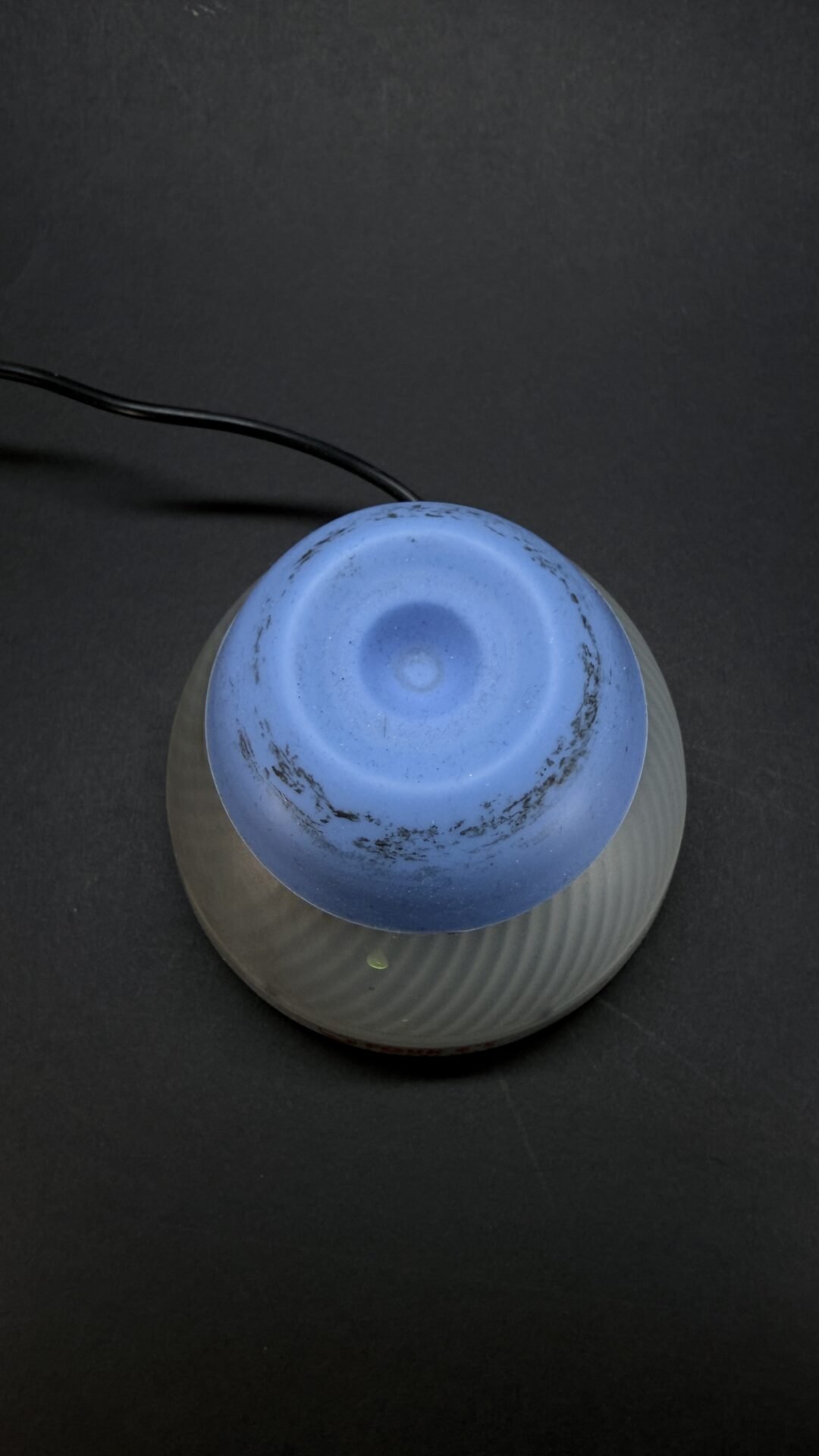 Overhead view of a blue ceramic, bowl-shaped lampshade atop a ribbed gray base, with a black cord extending from the left against a dark background.