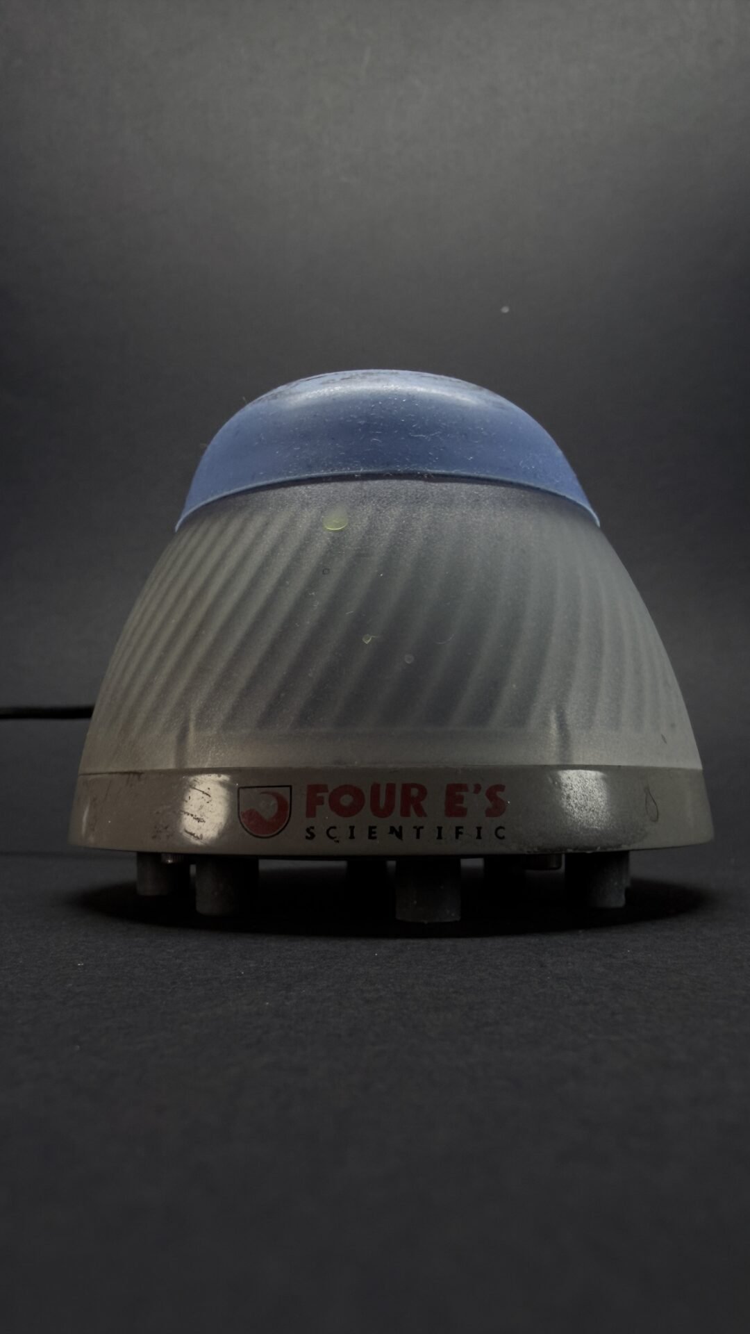 Close-up of a dome-shaped laboratory device with a blue top and ribbed gray body, branded 'FOUR E'S SCIENTIFIC' on a dark surface.