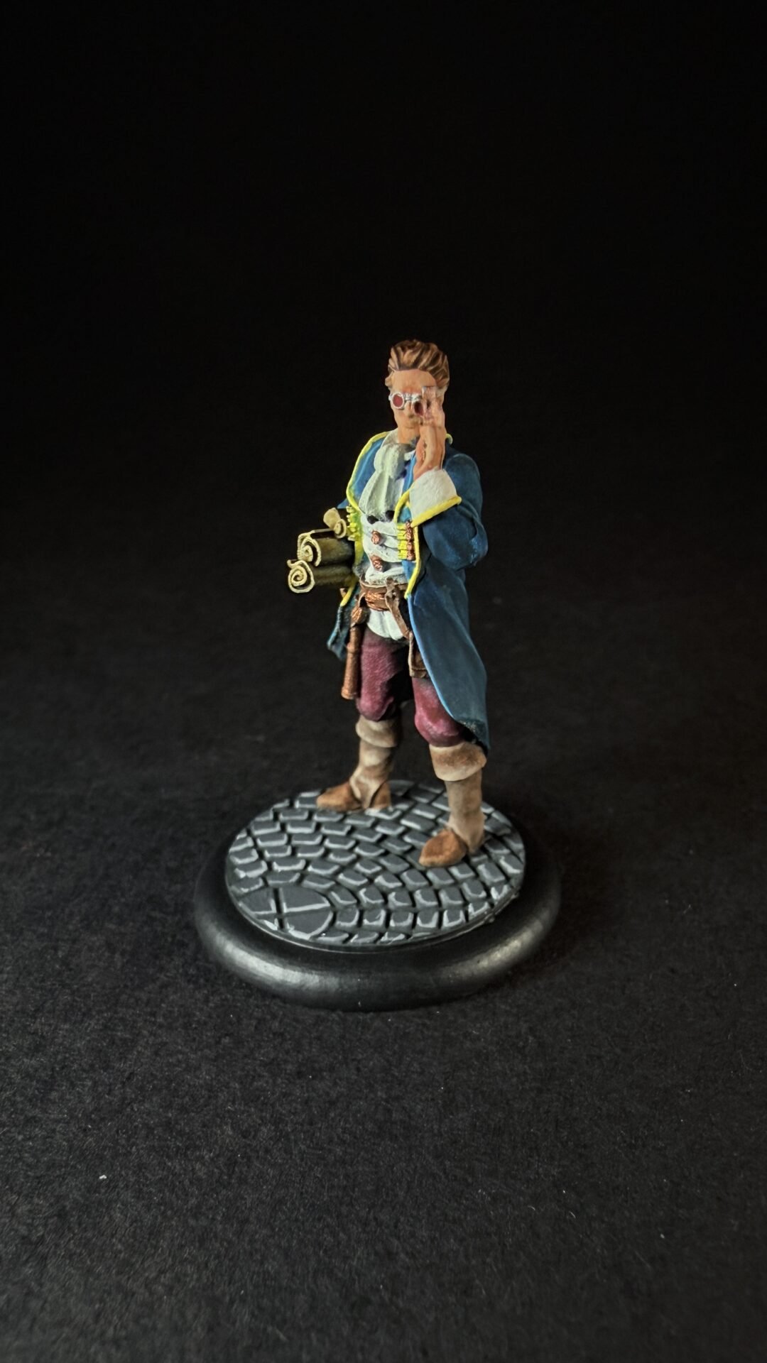 Miniature fantasy explorer figure wearing a blue coat with yellow trim, red pants, and boots, holding a small telescope and standing on a textured cobblestone base.