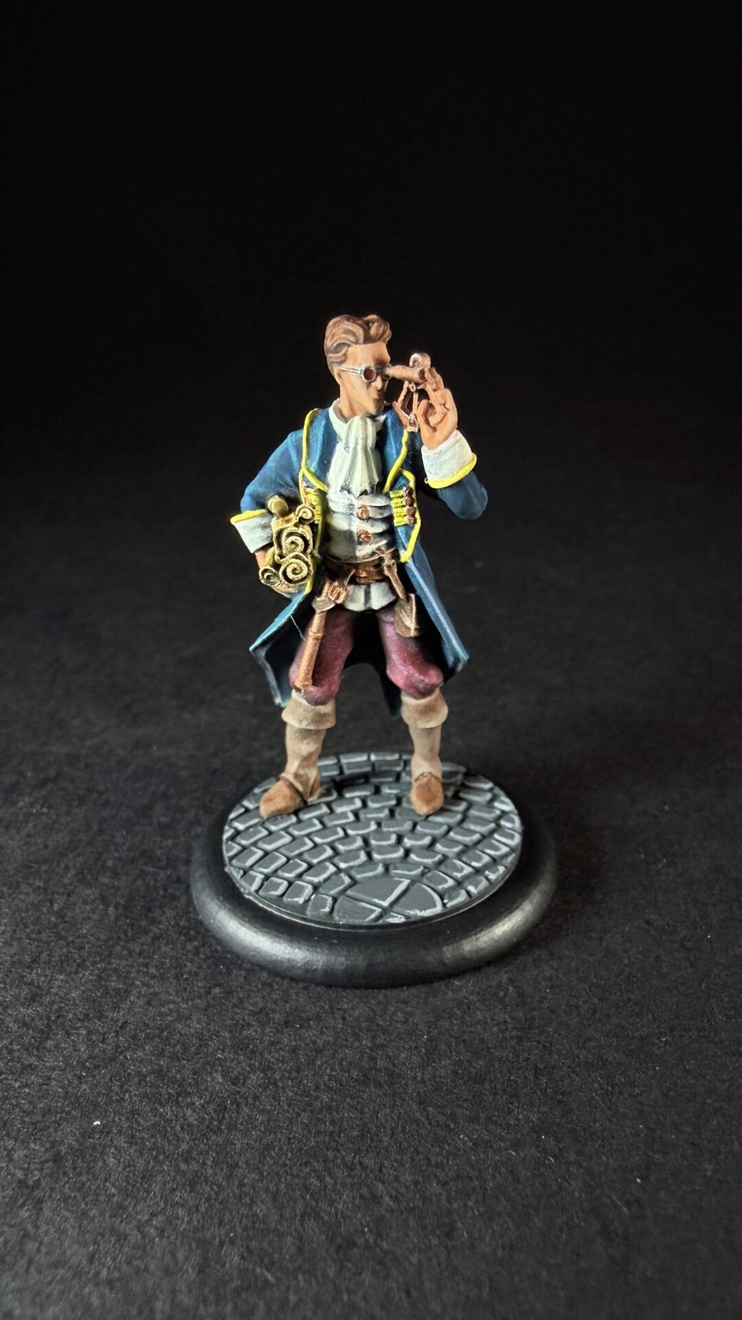 Painted fantasy mini figure: a man in a blue coat with yellow trim, white cravat, and brown boots, looking through a brass telescope on a cobblestone base.
