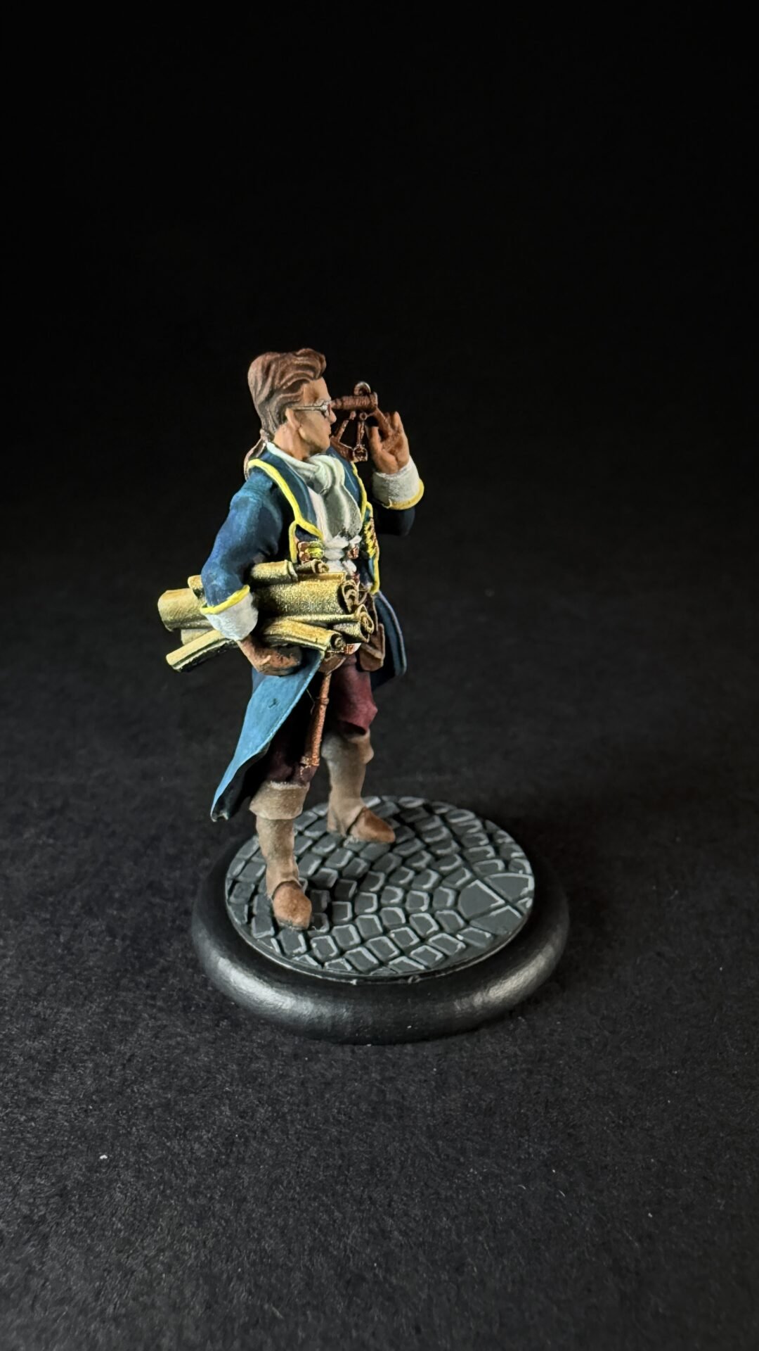 Miniature explorer in a blue coat with a telescope to the eye, carrying rolled parchments, standing on a cobblestone base.