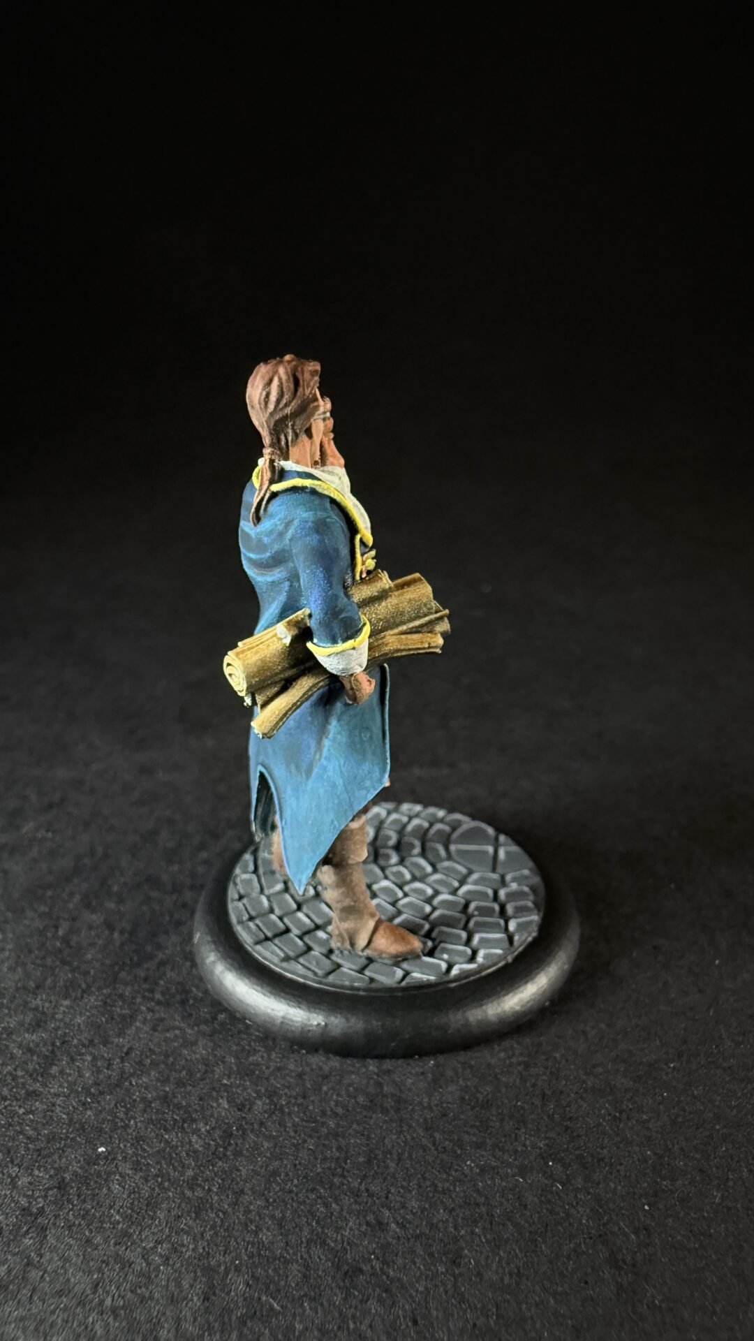 Miniature figure in a blue coat holding rolled scrolls, standing on a cobblestone base, facing left against a dark background.