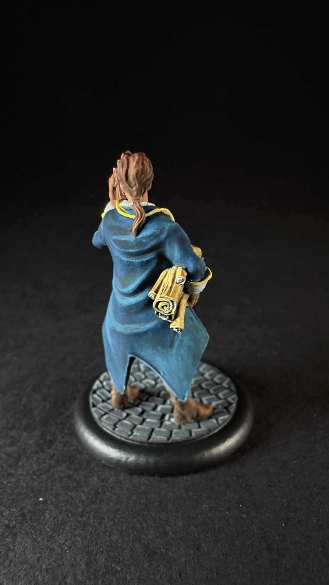 Back view of a miniature figure in a blue cloak holding rolled parchments, standing on a cobblestone base.