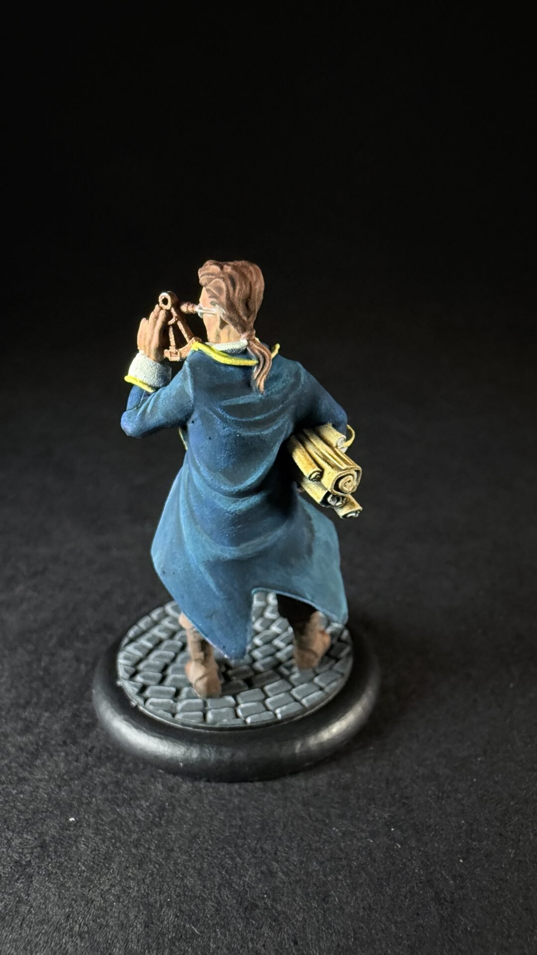 Miniature explorer in a blue coat holds a telescope to his eye, with rolled maps tucked under his arm, standing on a cobblestone base.
