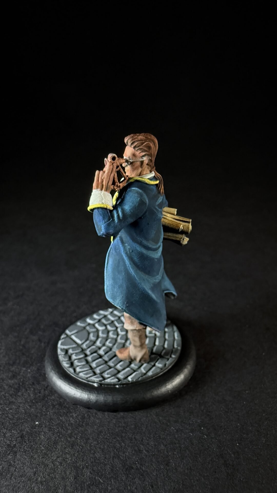 Figurine of a person in a blue coat peering through a telescope, with a bundle of wood on their back, standing on a cobblestone base.
