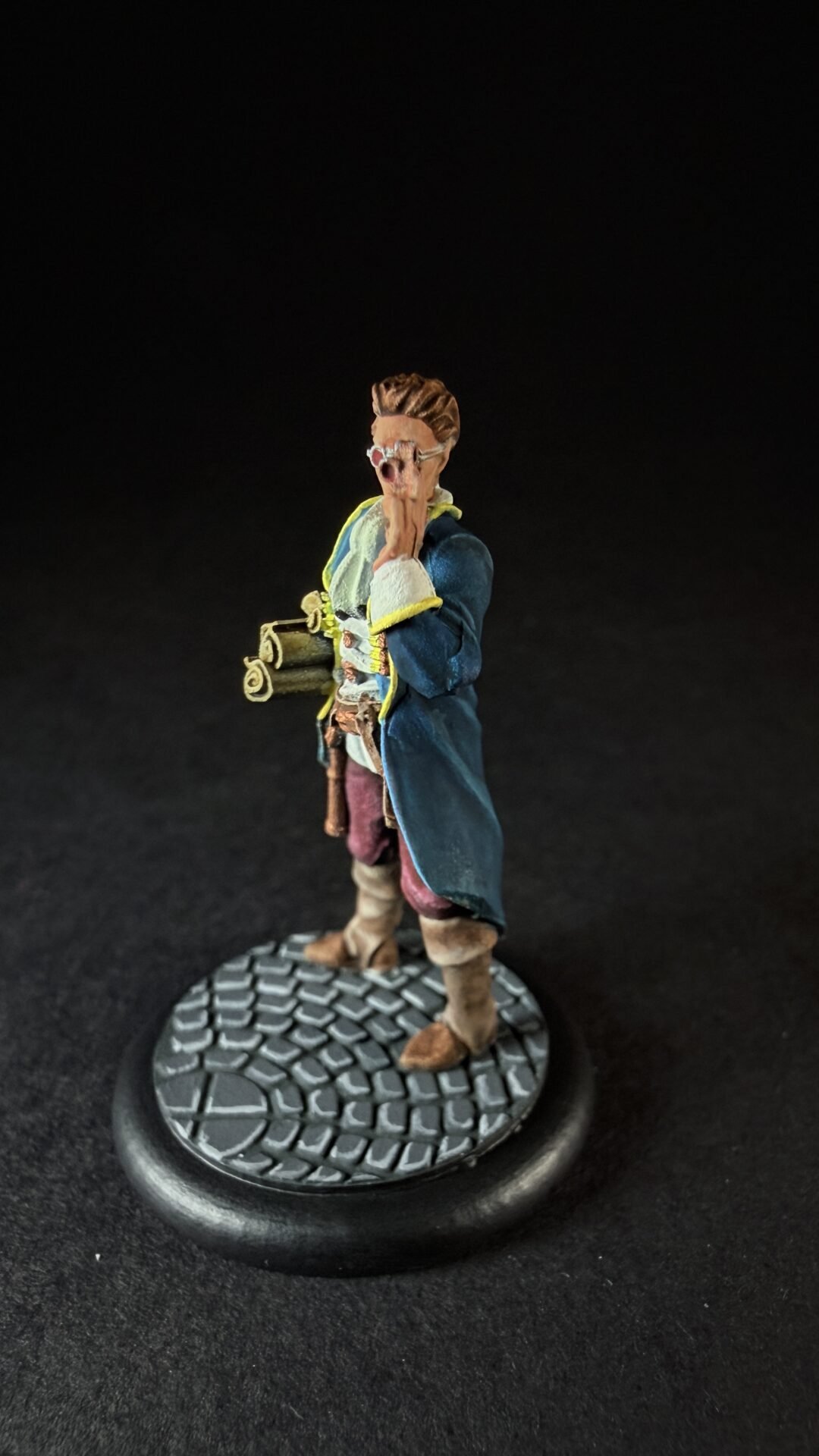 Miniature male explorer figure in a blue coat with yellow trim, standing on a cobblestone base, shouting with hands near his mouth and a rolled map under his arm.