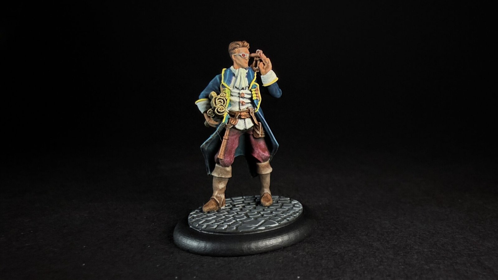 Miniature pirate captain in a blue coat with yellow trim, holding a telescope to his eye, standing on a textured circular base.