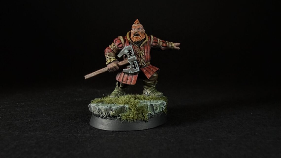Miniature dwarf warrior in red armor wielding a double-headed axe on a rocky grassy base, black background.