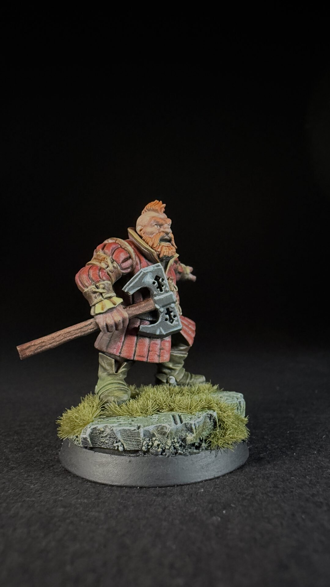 Miniature dwarf warrior in red armor, gripping a double-headed axe, standing on a grassy rocky base.