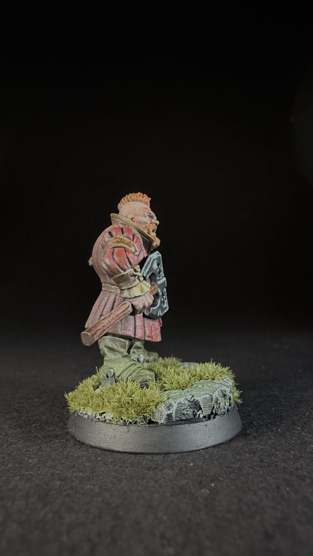 Painted fantasy miniature of a bearded warrior in red armor, carrying a shield and club, standing on a mossy rocky base