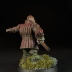 Back view of a miniature medieval knight in red segmented armor, standing on a grassy base with a sword in hand against a dark background, seen from behind.