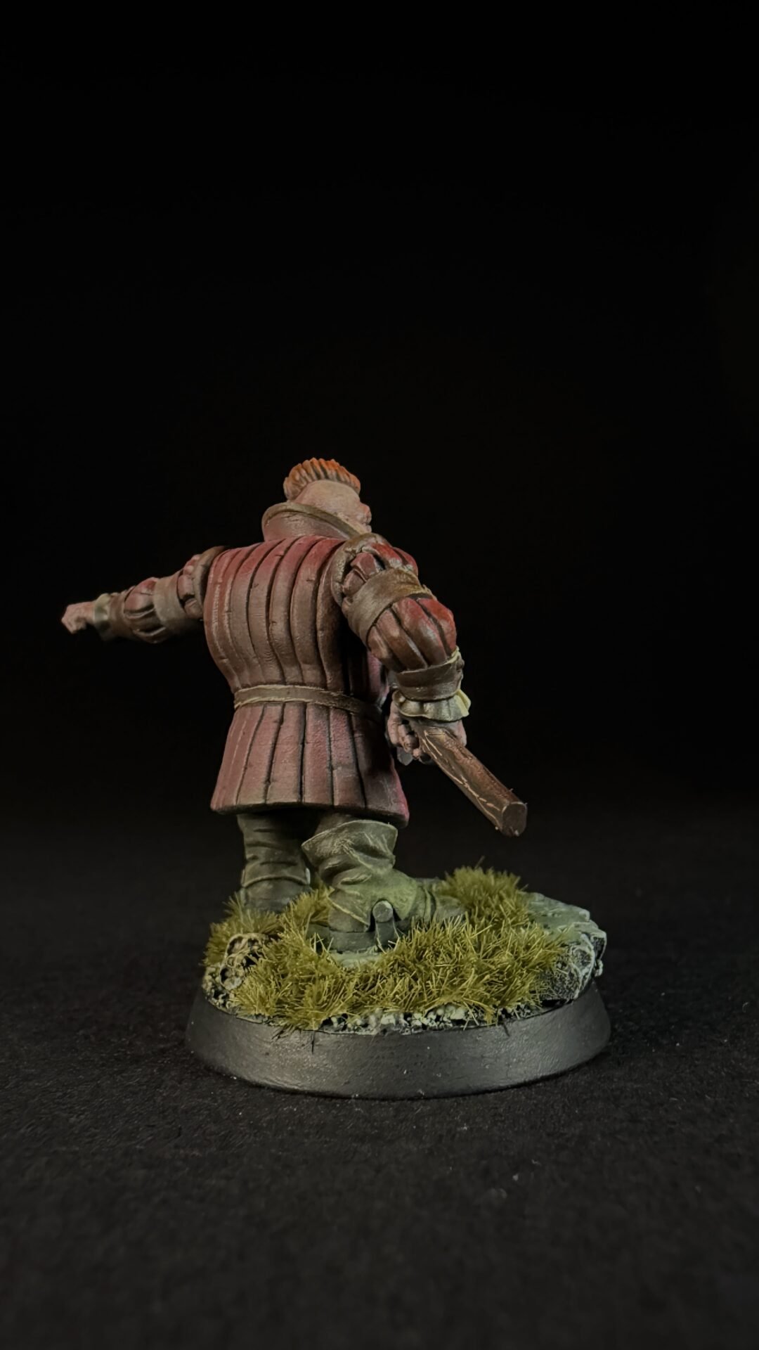 Back view of a miniature medieval knight in red segmented armor, standing on a grassy base with a sword in hand against a dark background, seen from behind.