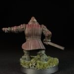 Back view of a red-brown armored knight miniature with a raised left arm and sword, standing on a grassy base with a skull on dark background