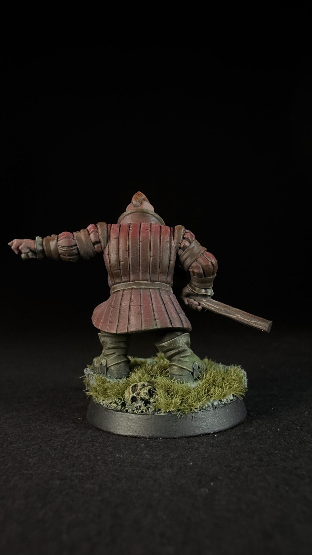 Back view of a red-brown armored knight miniature with a raised left arm and sword, standing on a grassy base with a skull on dark background