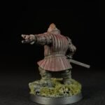 Back view of a medieval knight miniature in red armor, with right arm extended as if pointing or gesturing, sword at the hip, standing on a grassy base.