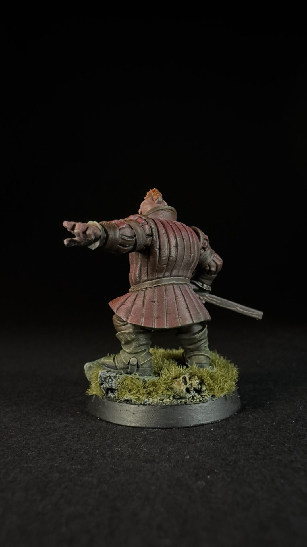 Back view of a medieval knight miniature in red armor, with right arm extended as if pointing or gesturing, sword at the hip, standing on a grassy base.