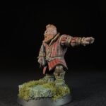 Fantasy dwarf miniature in red armor with beard, standing on a grassy rocky base, arm extended forward.