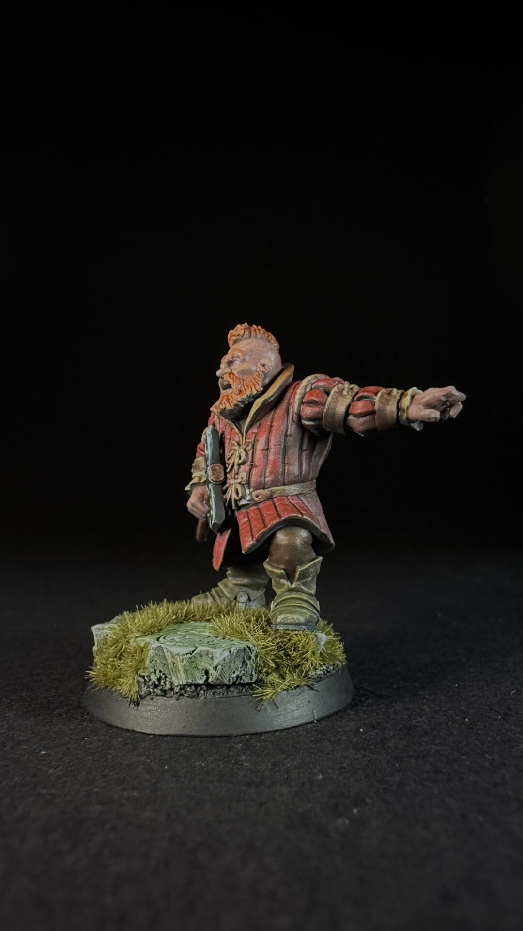 Fantasy dwarf miniature in red armor with beard, standing on a grassy rocky base, arm extended forward.