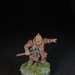 Dwarf warrior miniature in red armor, gripping a double-headed axe, standing on a grassy rocky base.