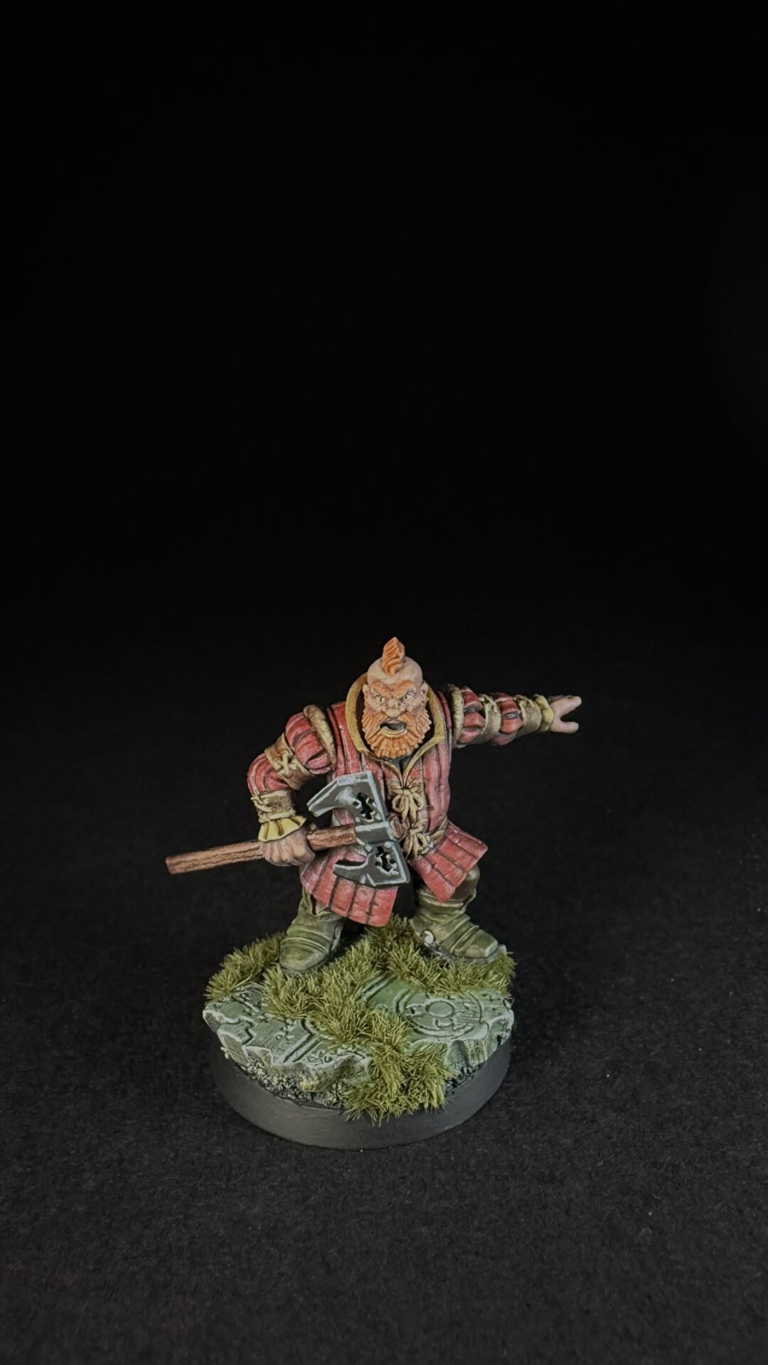 Dwarf warrior miniature in red armor, gripping a double-headed axe, standing on a grassy rocky base.