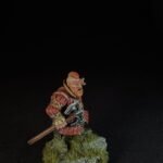 Miniature dwarf warrior with orange beard in red armor, holding a battle axe, standing on a mossy, rocky base.