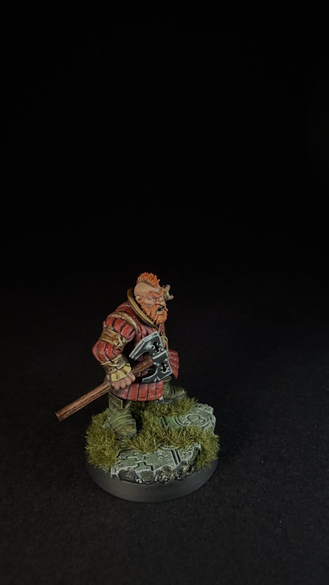 Miniature dwarf warrior with orange beard in red armor, holding a battle axe, standing on a mossy, rocky base.