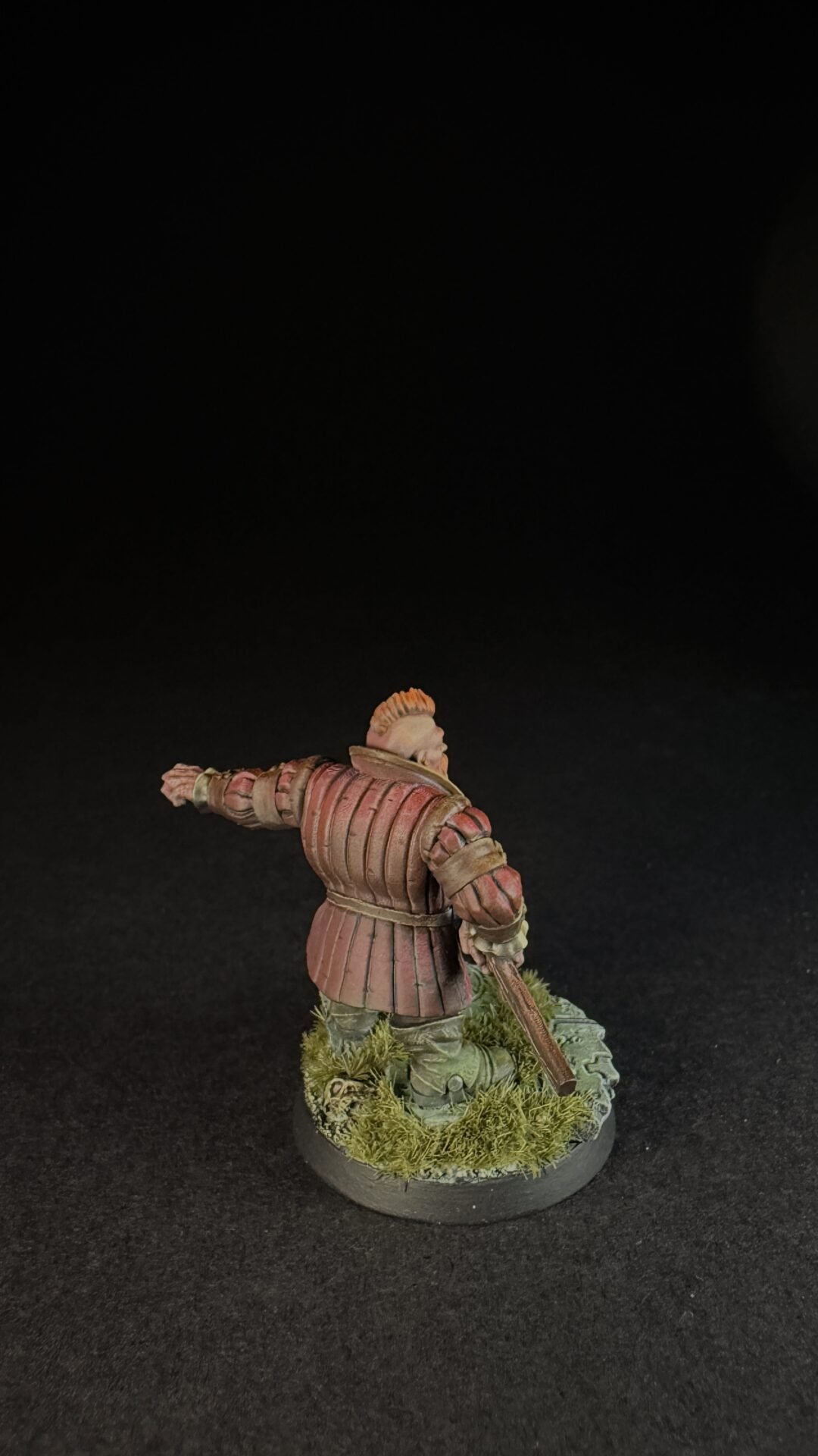 Back view of a medieval knight miniature in red-brown plate armor, standing on a grassy base with a sword sheathed at the side.