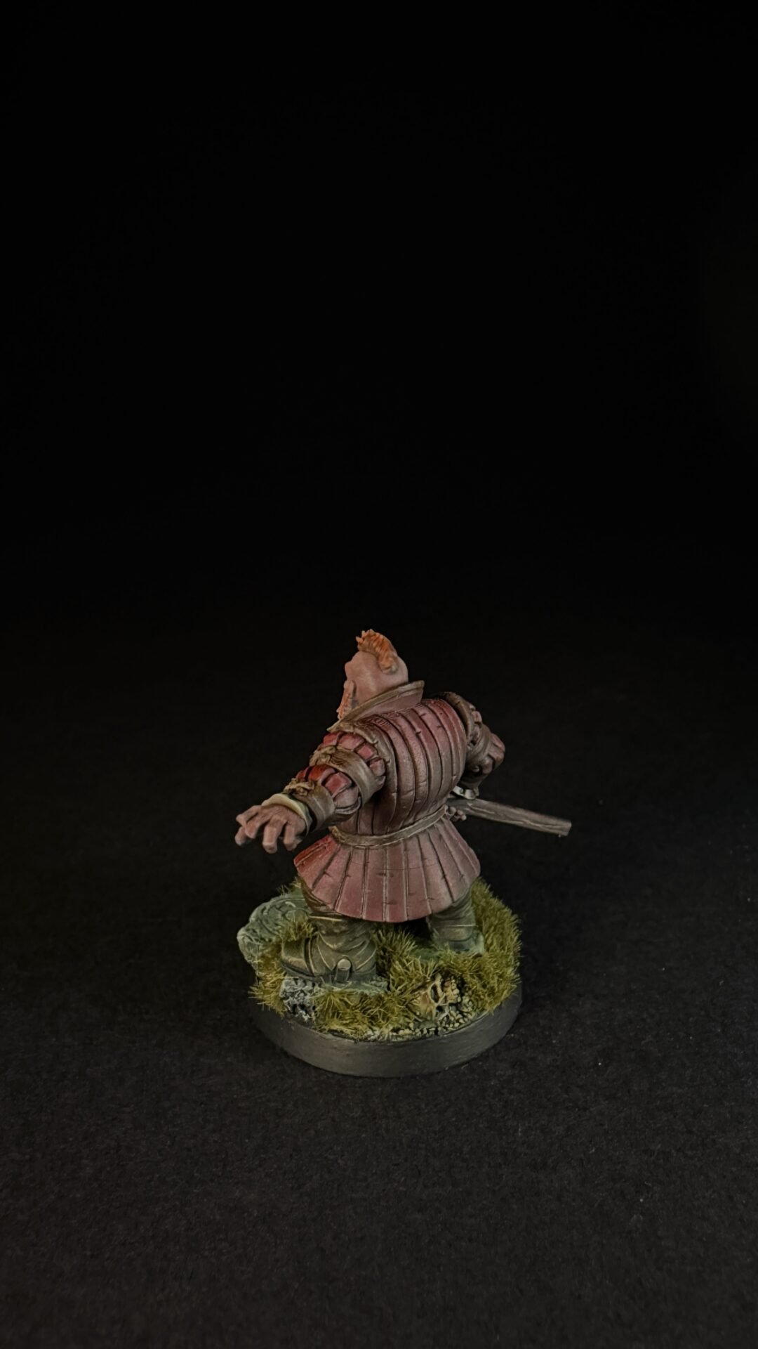 Back view of a medieval knight miniature in pinkish armor, with a sword at the side, standing on a grassy base.