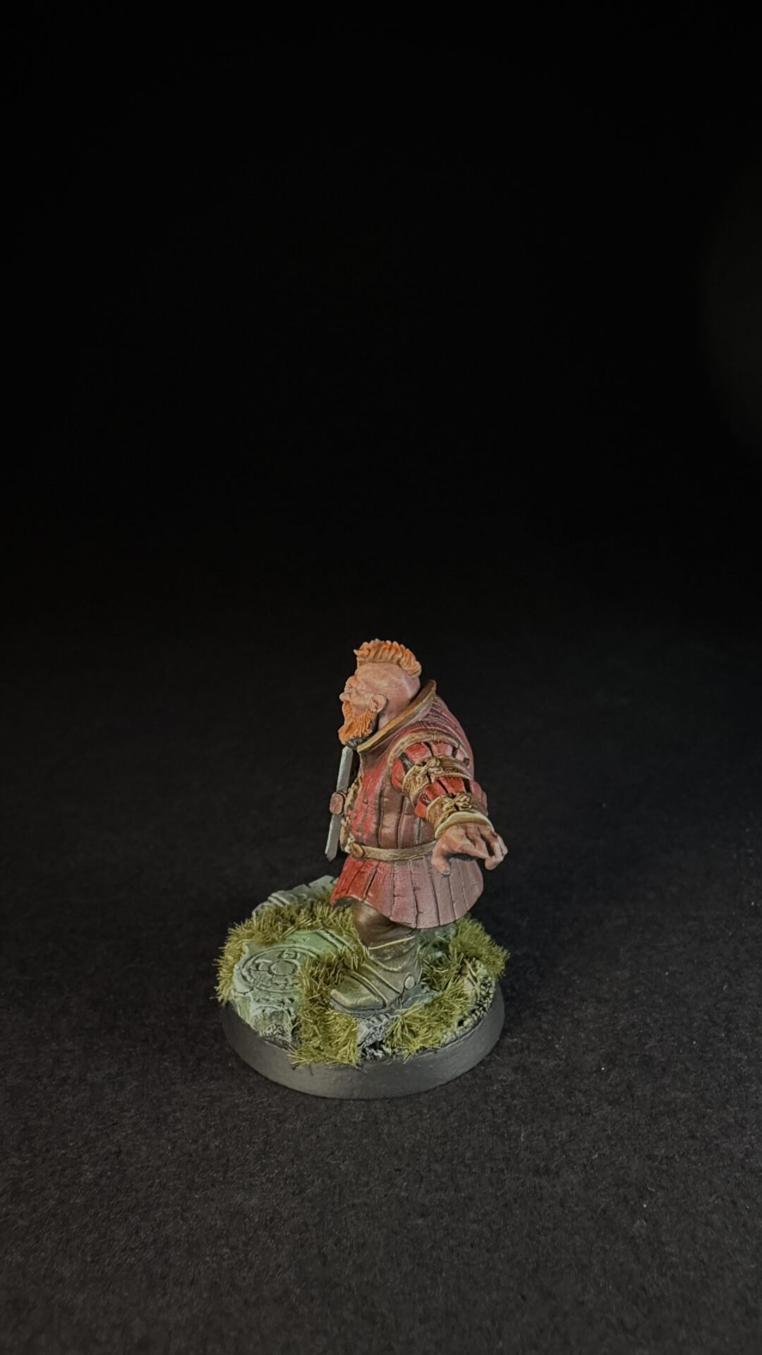 Miniature bearded warrior in red armor standing on a mossy base against a black background.