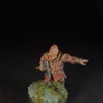 Fantasy dwarf miniature in red armor, wielding a war hammer with an outstretched left arm, standing on a grassy stone base.
