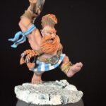 Fantasy dwarf warrior miniature with orange beard, hammer raised, blue scarf, and striped belt standing on a rocky base.