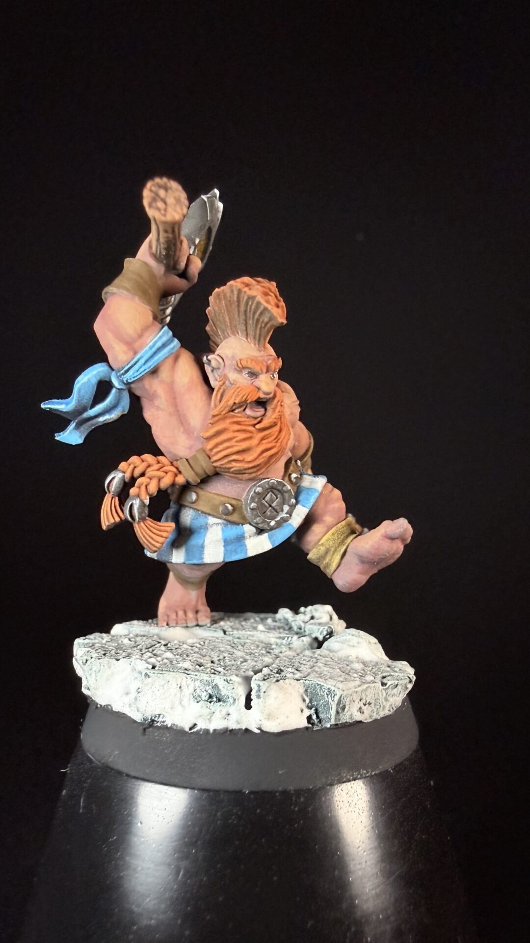 Fantasy dwarf warrior miniature with orange beard, hammer raised, blue scarf, and striped belt standing on a rocky base.
