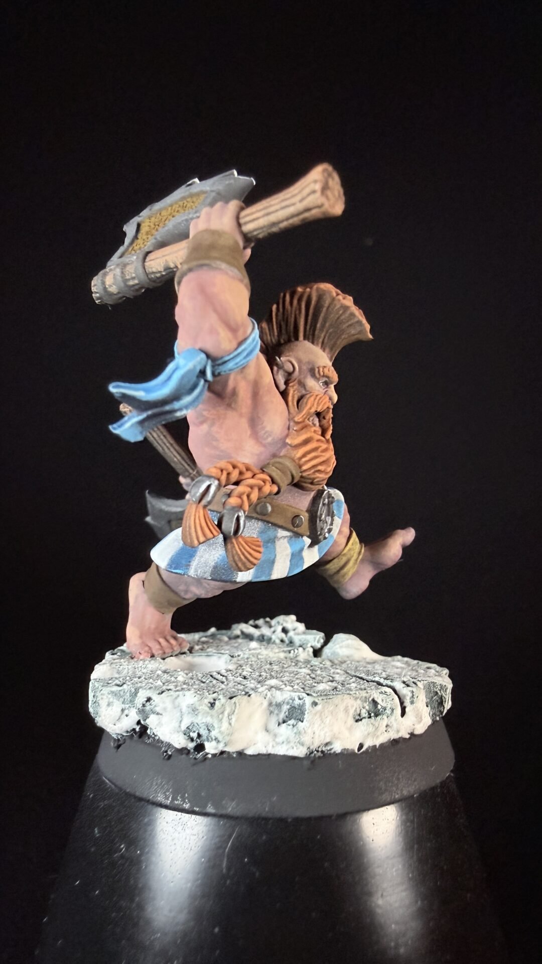 Fantasy dwarf warrior miniature, bare-ch chest, braided beard, blue scarf, striped kilt, and a large hammer raised as if mid-swing on a rocky base