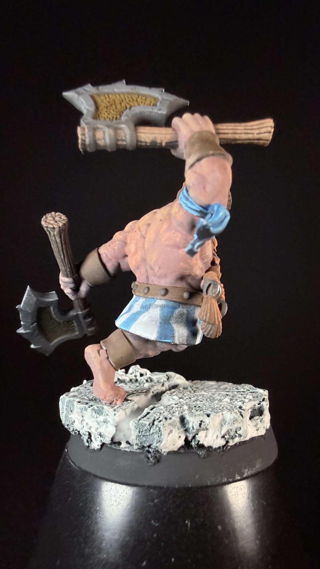 Miniature fantasy barbarian figure mid-stride, wielding a large double-headed axe and a wooden club, wearing a blue-striped kilt and tan armor, standing on a rocky base.