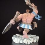 Painted fantasy miniature of a muscular pink-skinned warrior swinging a large bladed club, wearing a blue-striped kilt and scarf on a rocky base.