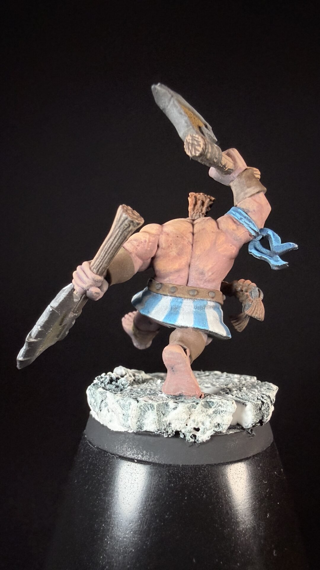 Painted fantasy miniature of a muscular pink-skinned warrior swinging a large bladed club, wearing a blue-striped kilt and scarf on a rocky base.