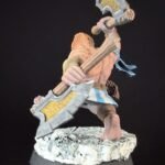 Fantasy miniature figure: a muscular warrior wielding a large double-headed war axe, standing on a rocky base with a blue sash fluttering behind him.