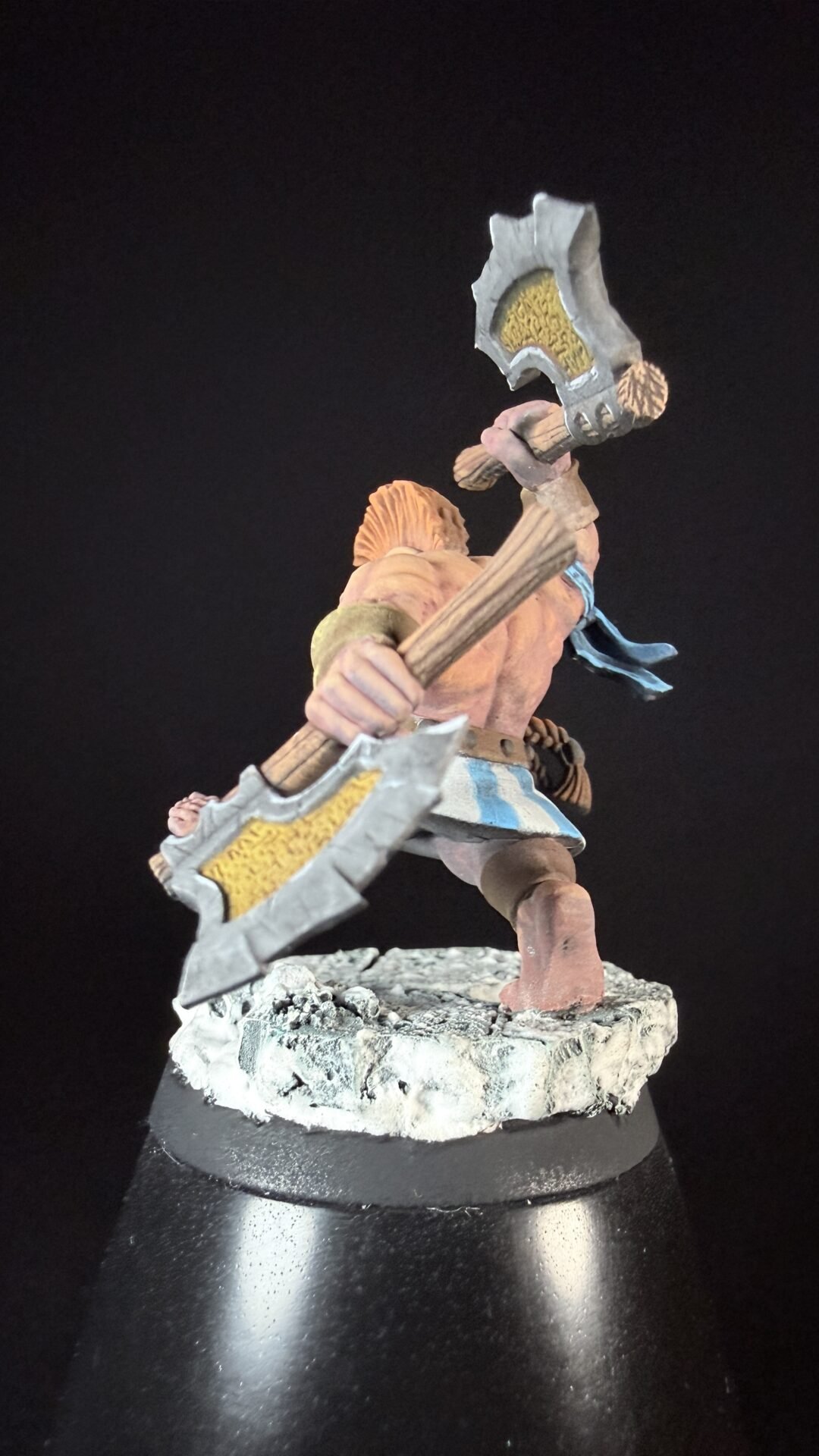Fantasy miniature figure: a muscular warrior wielding a large double-headed war axe, standing on a rocky base with a blue sash fluttering behind him.