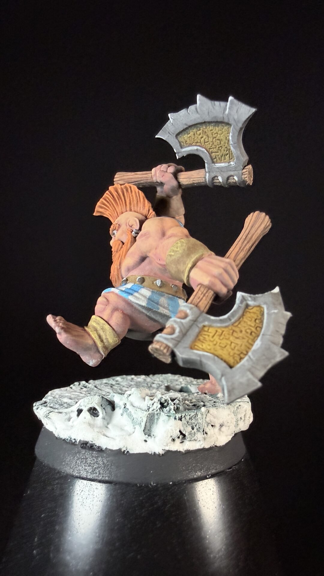 Miniature fantasy warrior with two axes, orange beard, bare chest, blue-striped loincloth, standing on a rocky base.