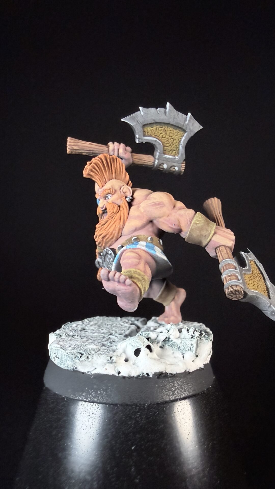 Tabletop fantasy dwarf miniature in a leaping battle pose, wielding a large axe over his shoulder and a hammer in his other hand, standing on a rocky snowy base.