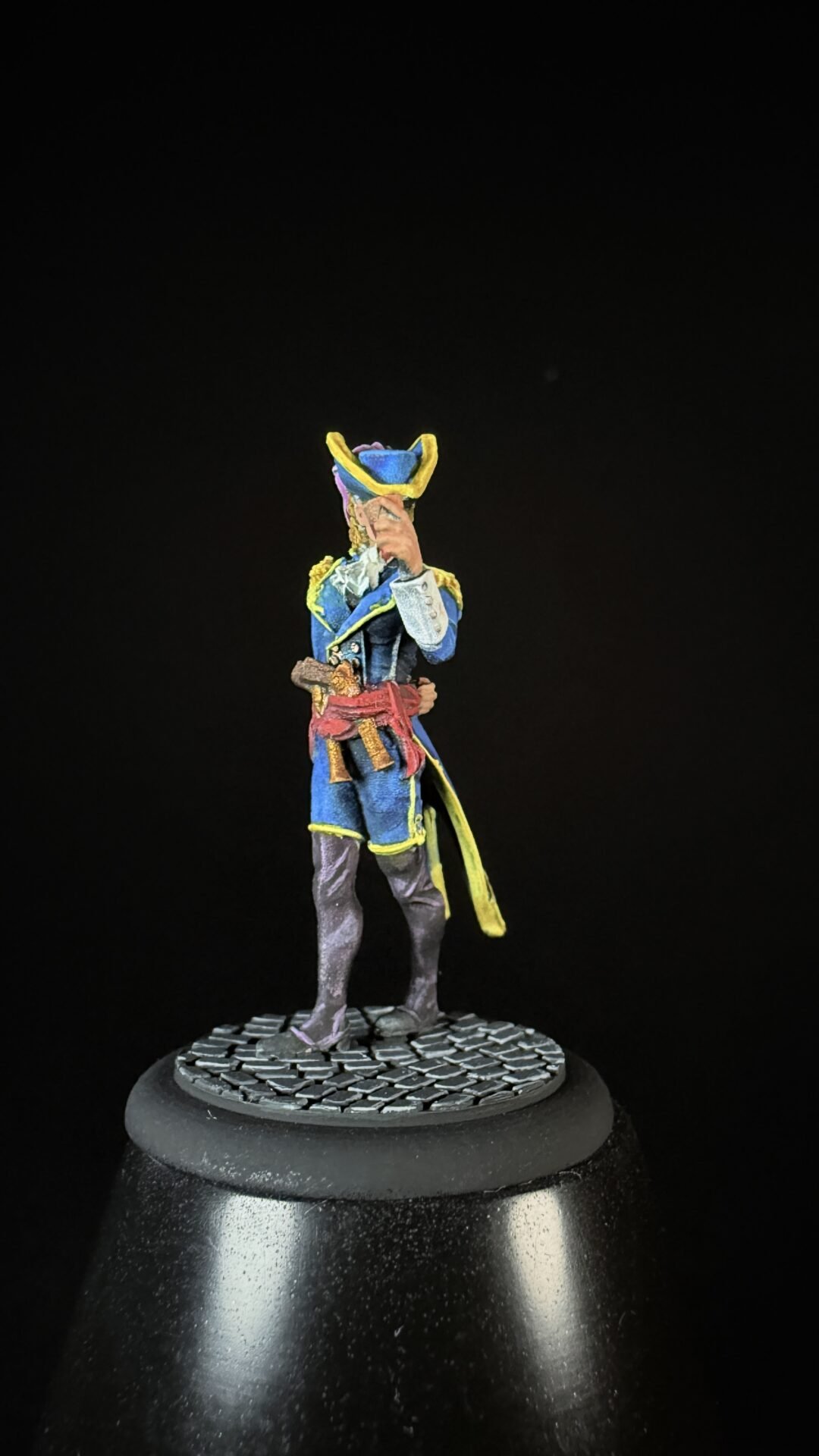 Miniature pirate captain in a blue coat with yellow trim, holding a pistol in his right hand, standing on a cobblestone base on a black background.