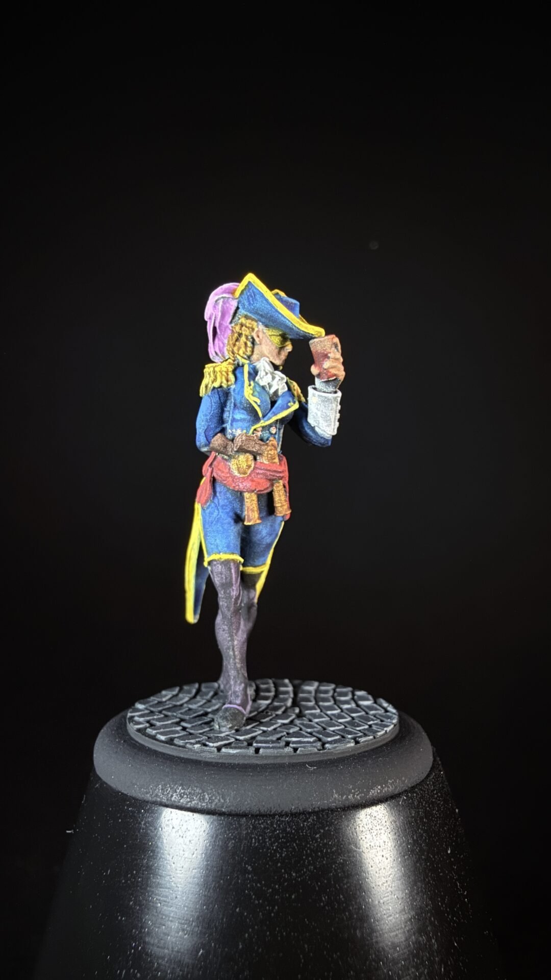Miniature pirate figure in a blue coat with yellow trim and a tricorn hat, standing on a textured base against a black background