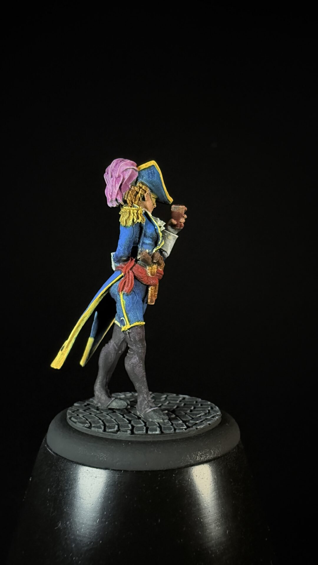 Miniature pirate figure in blue coat with yellow trim, pink scarf, holding a mug, on a cobblestone base.