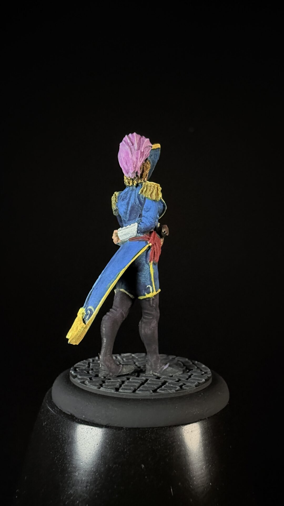 Small painted miniature of a soldier in a blue coat with yellow trim and a pink plume, standing on a textured cobblestone base.
