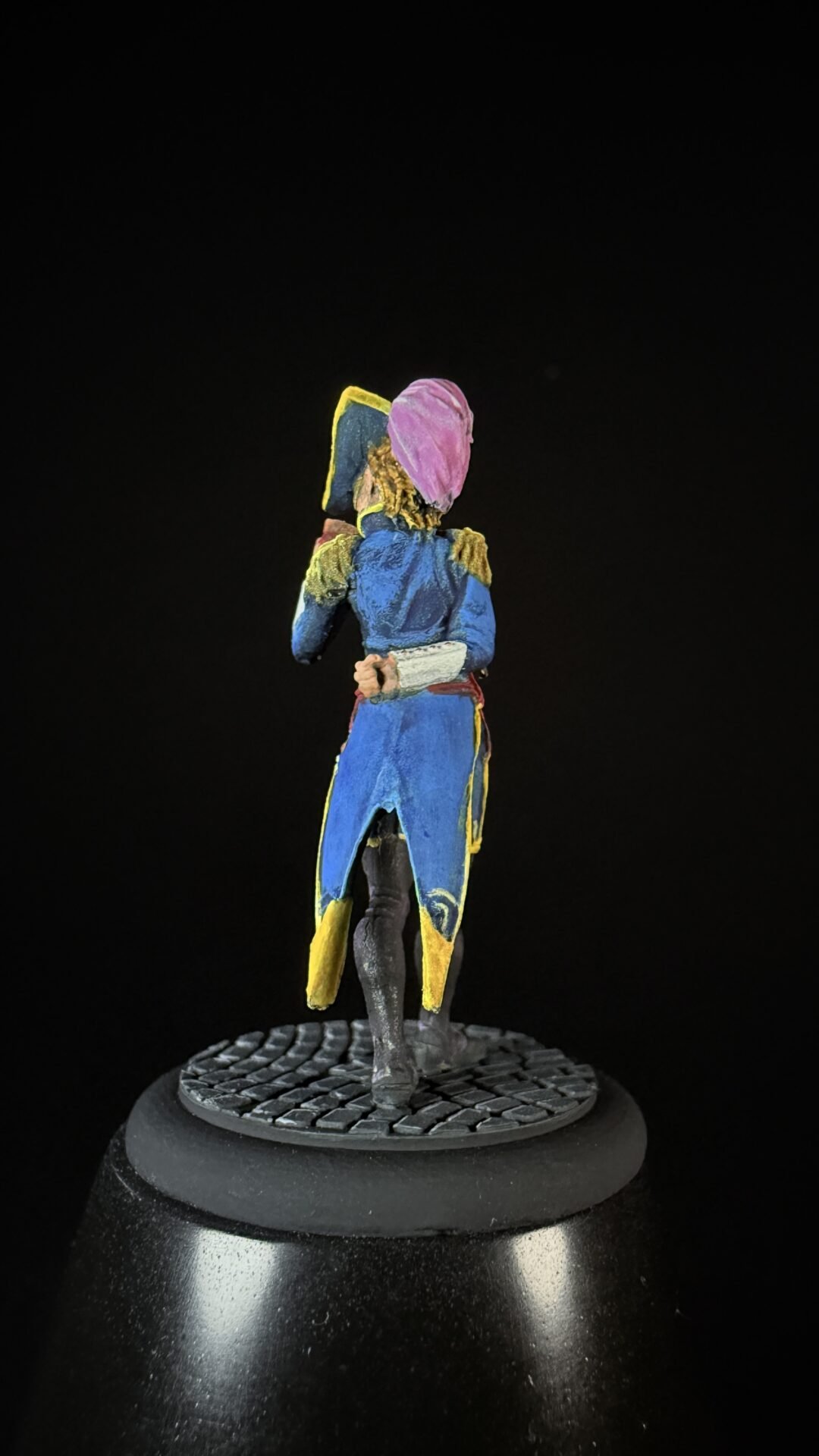 Back view of a small painted miniature figure in a blue coat with yellow trim and pink hood, standing on a textured black base.