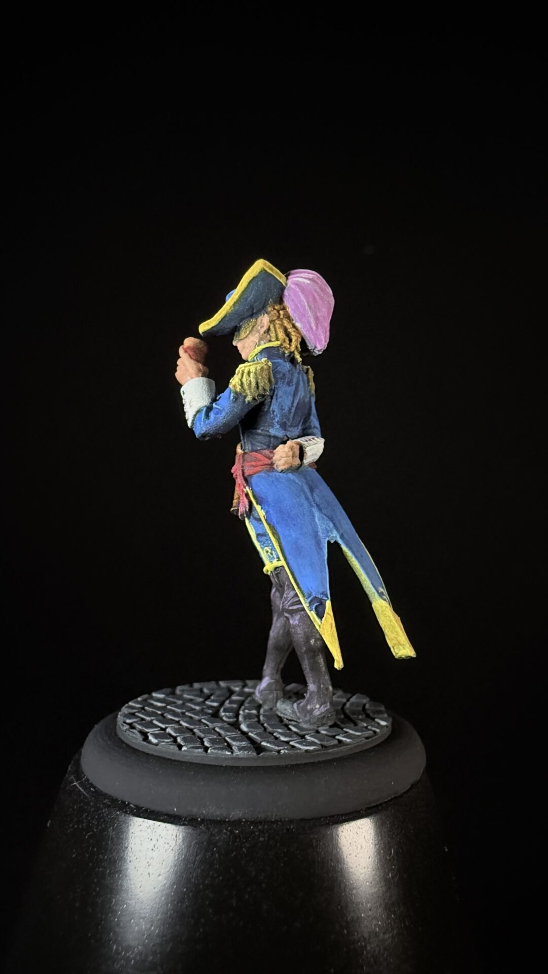 Colorful painted pirate miniature in a blue coat with yellow trim and a purple hat, standing on a textured base on a black background.