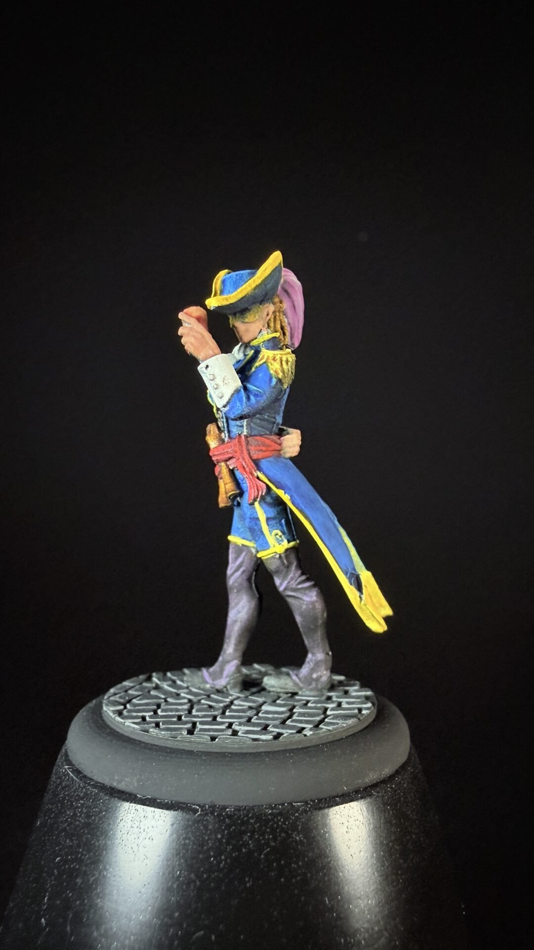 Miniature pirate/naval officer figure in a blue coat with yellow trim, red sash, and hat, standing on a textured base against a dark background.
